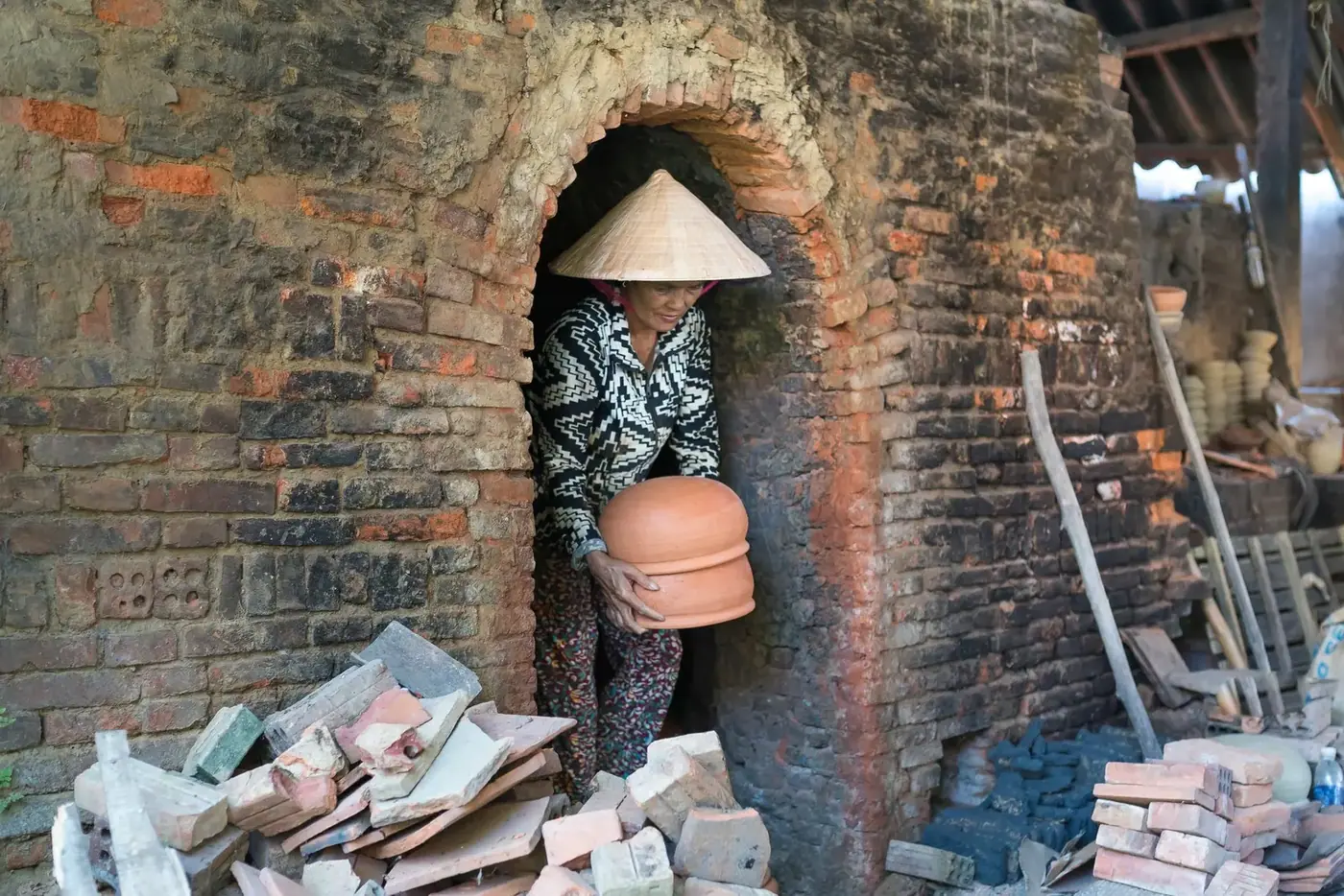 Woman in a conical hat shaping clay beside a traditional brick kiln in a Bat Trang alley