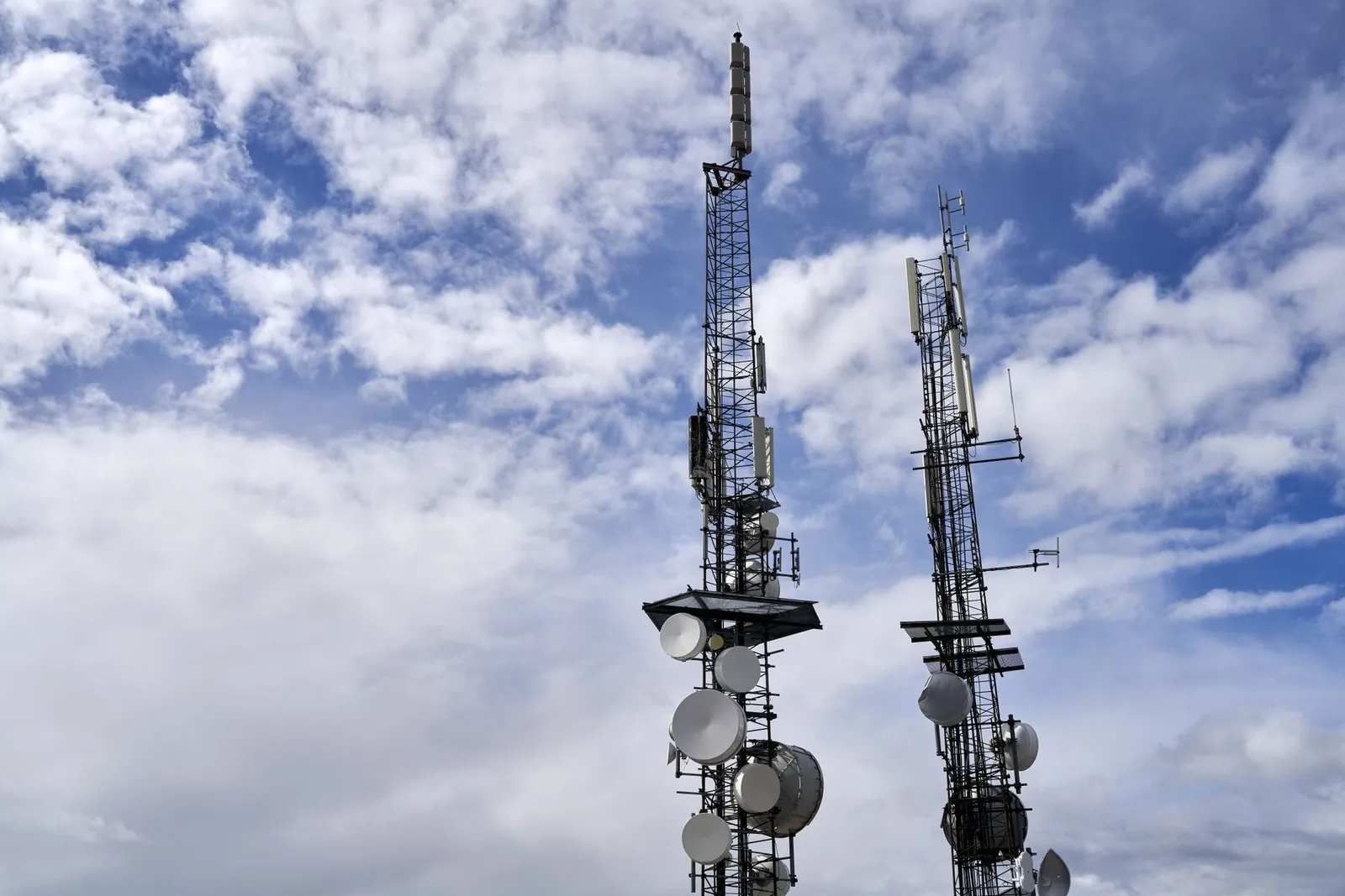 Two telecom towers with antennas and dishes against a cloudy blue sky