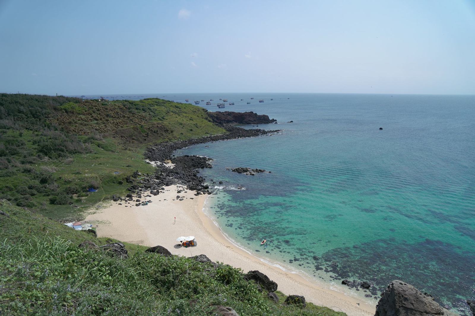 Green cliffs above a sandy cove with turquoise water and fishing boats offshore