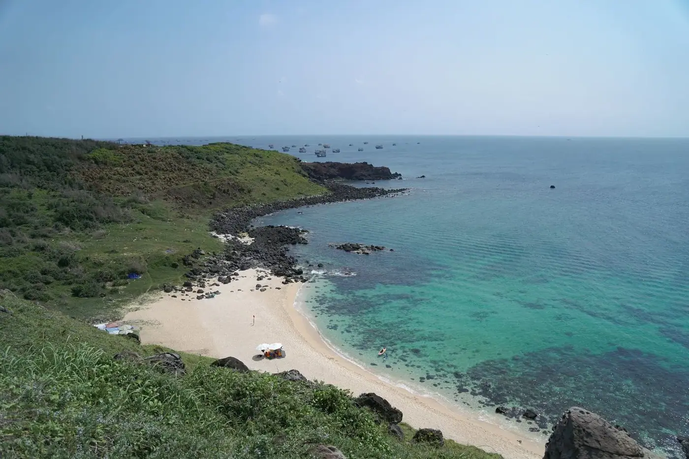 Green cliffs above a sandy cove with turquoise water and fishing boats offshore