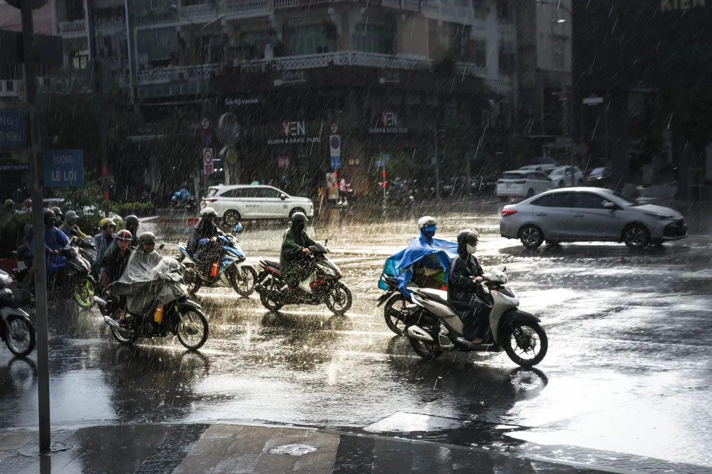 Motorbike riders in rain ponchos crossing a wet street in Ho Chi Minh City