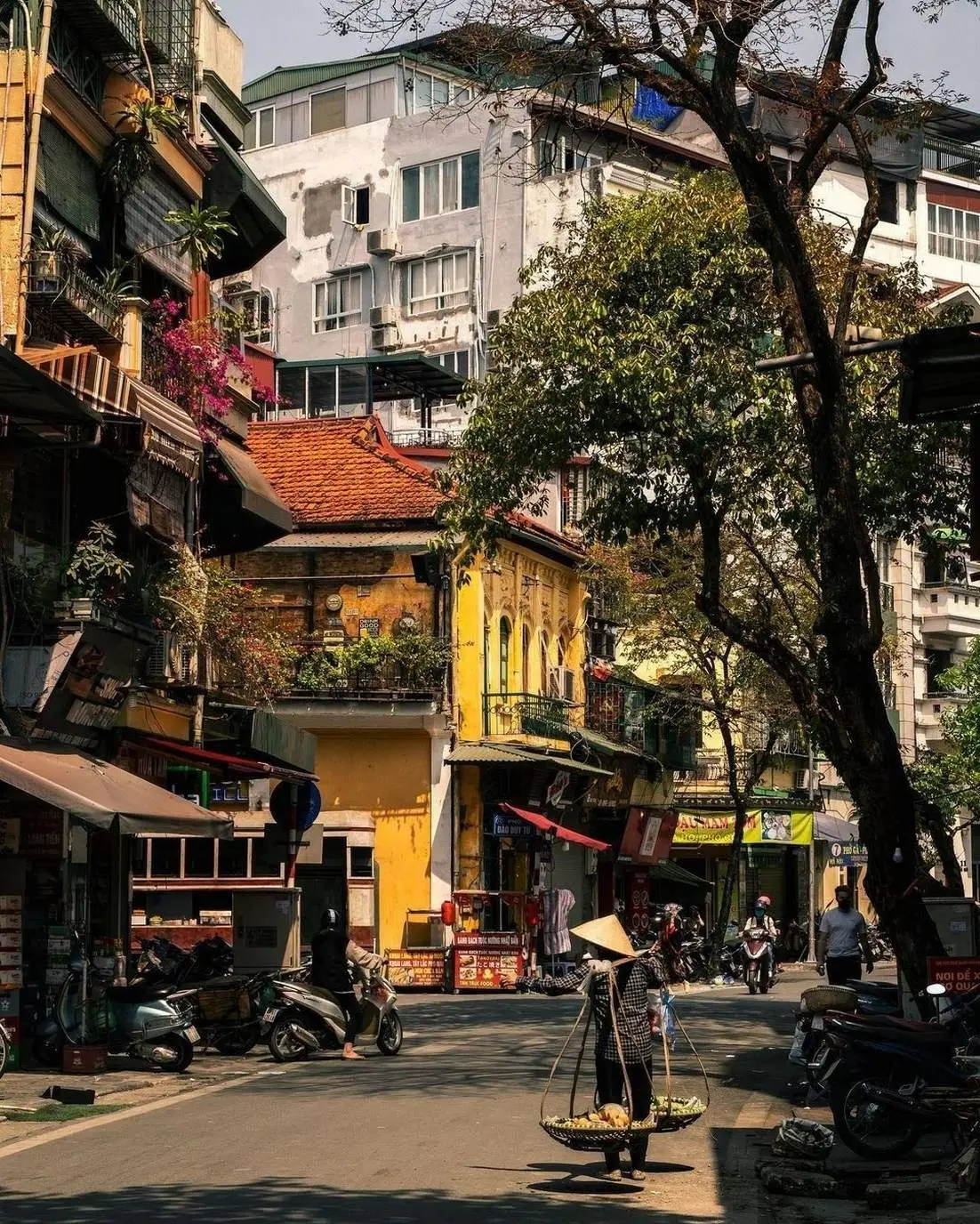 Street vendor carrying baskets on a pole past colorful old-quarter buildings in Hanoi