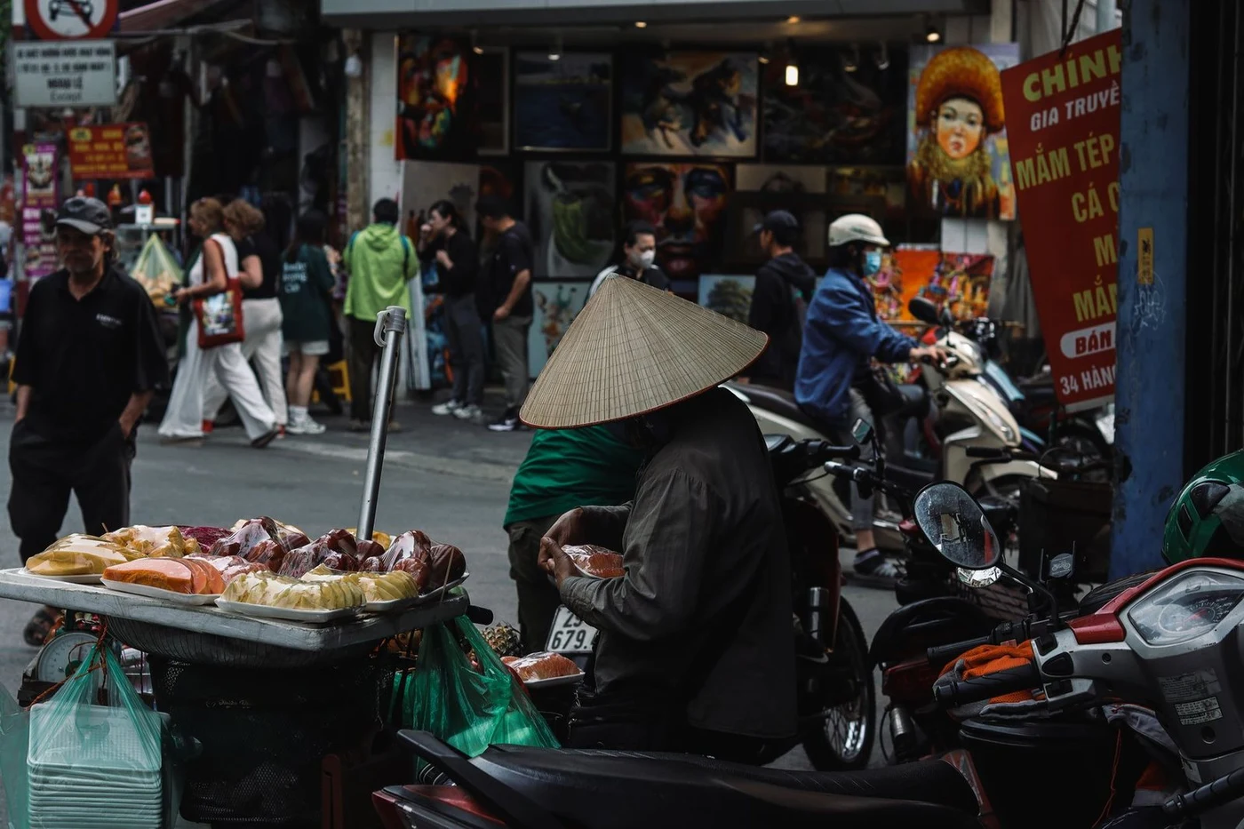 Street vendor in a conical hat sells sliced fruit beside parked scooters