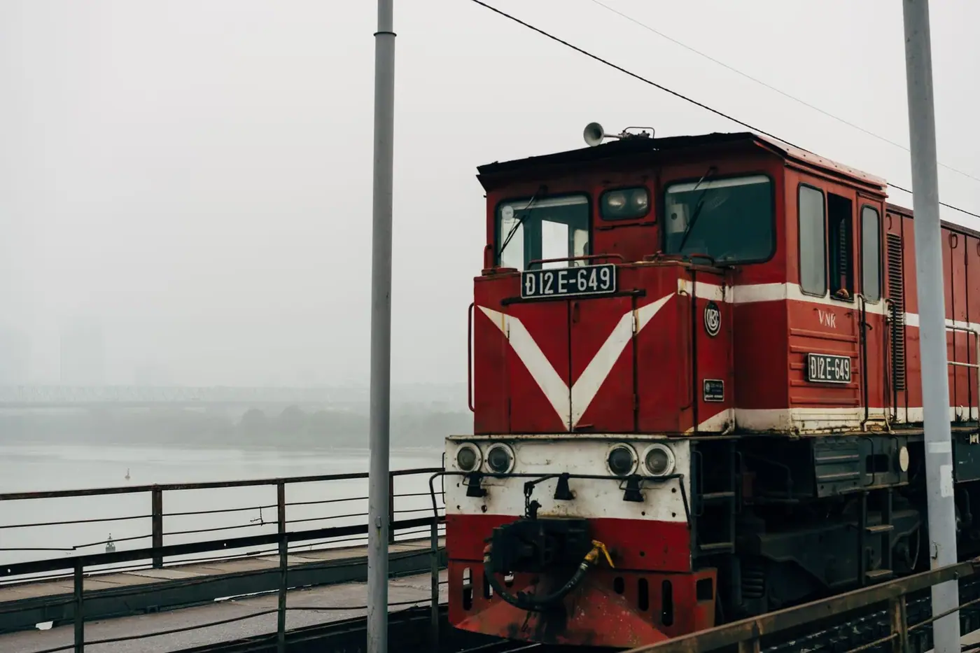 A red Vietnam Railways locomotive crossing a misty bridge into Hanoi