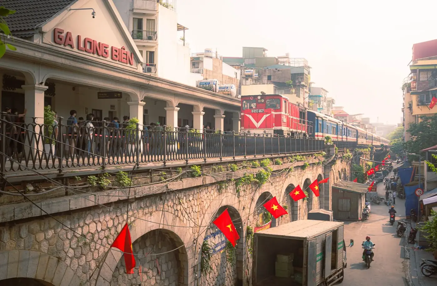 A train arriving at Ga Long Bien station in Hanoi with waiting passengers and Vietnamese flags