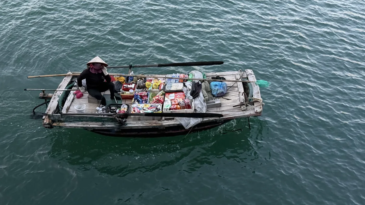 Vendor sitting on a wooden boat loaded with drinks and snacks on green water