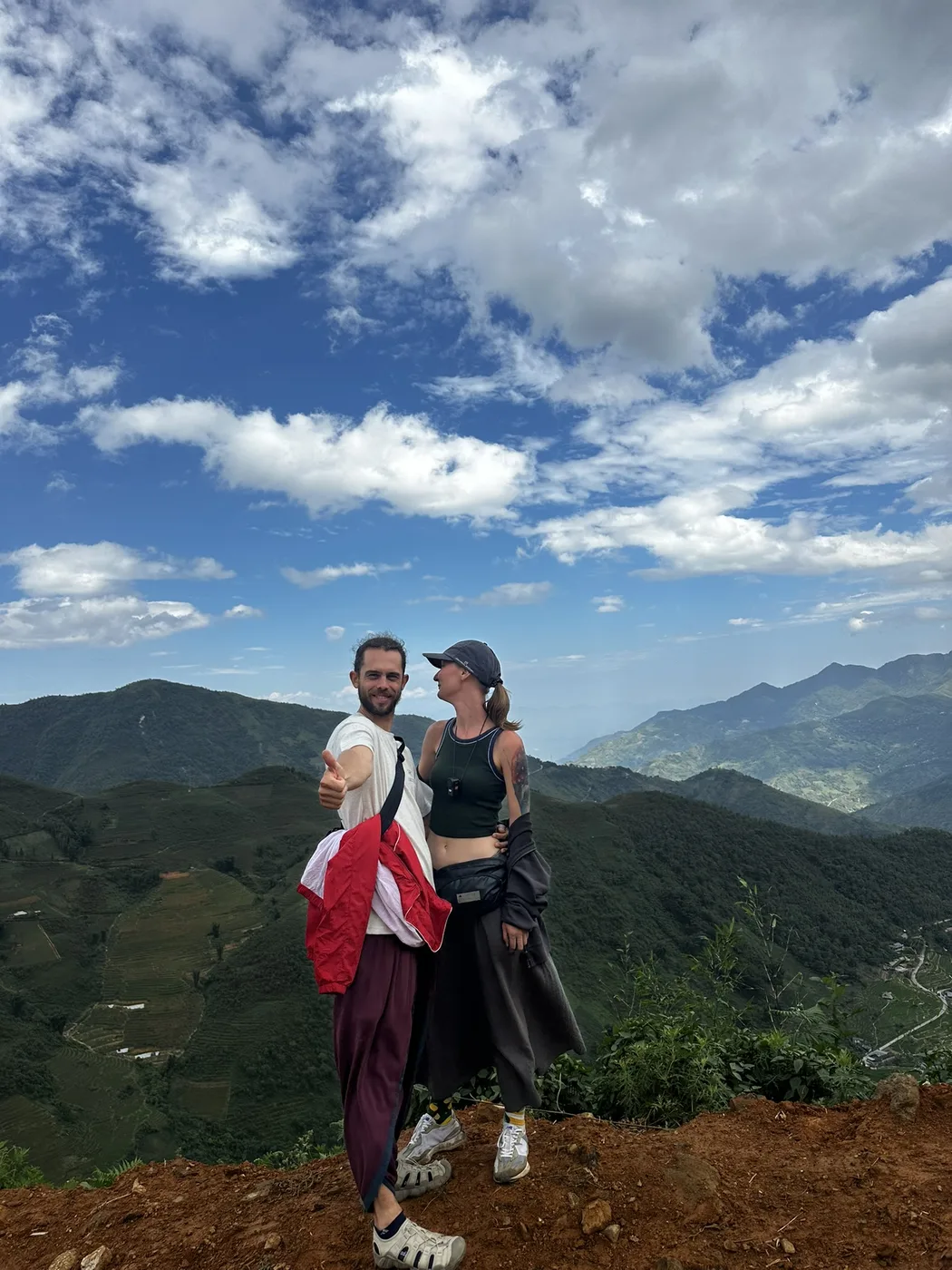 A couple stands smiling on a green mountain ridge under a blue sky with scattered clouds