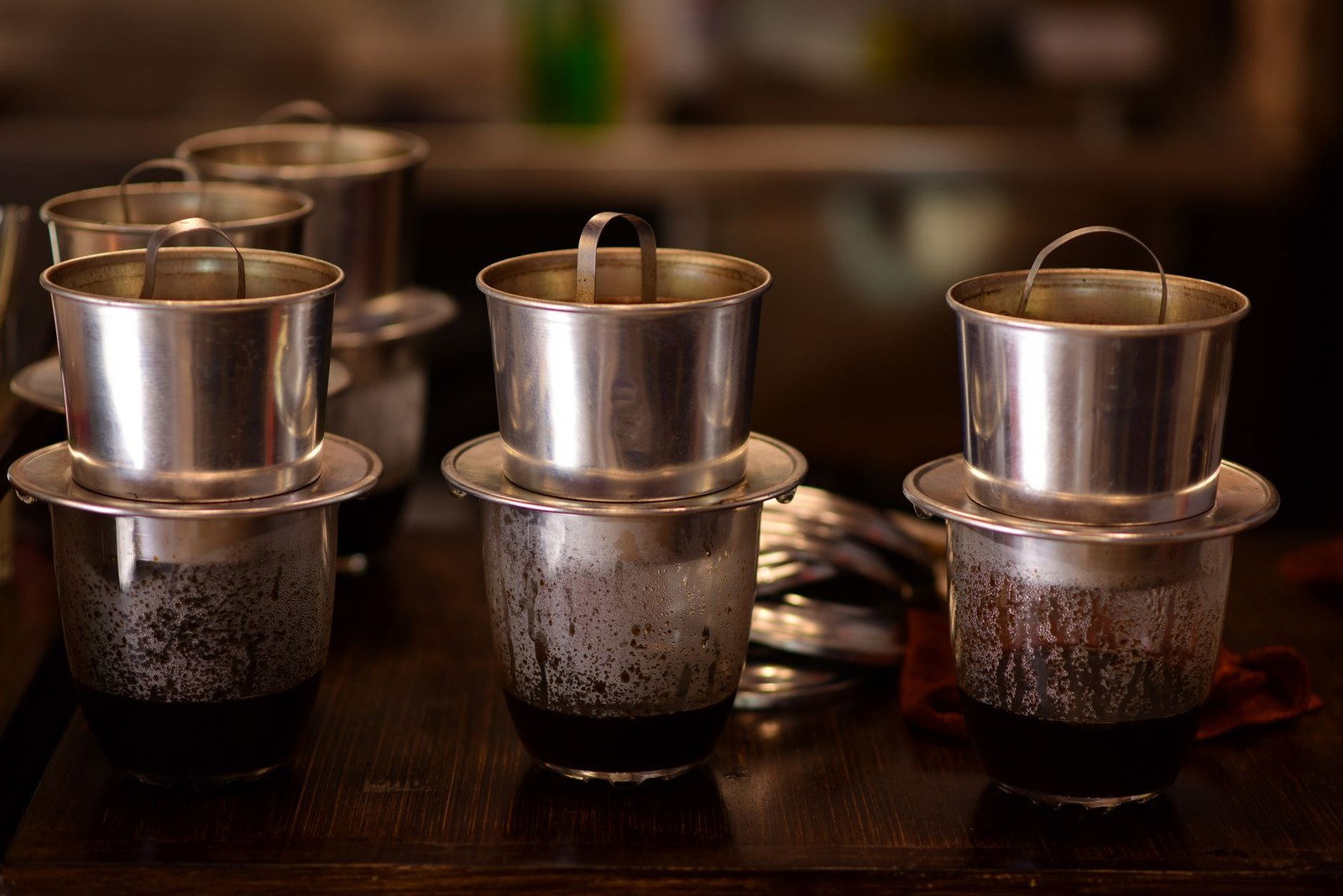 Several metal phin filters actively dripping coffee into glasses on a wooden table