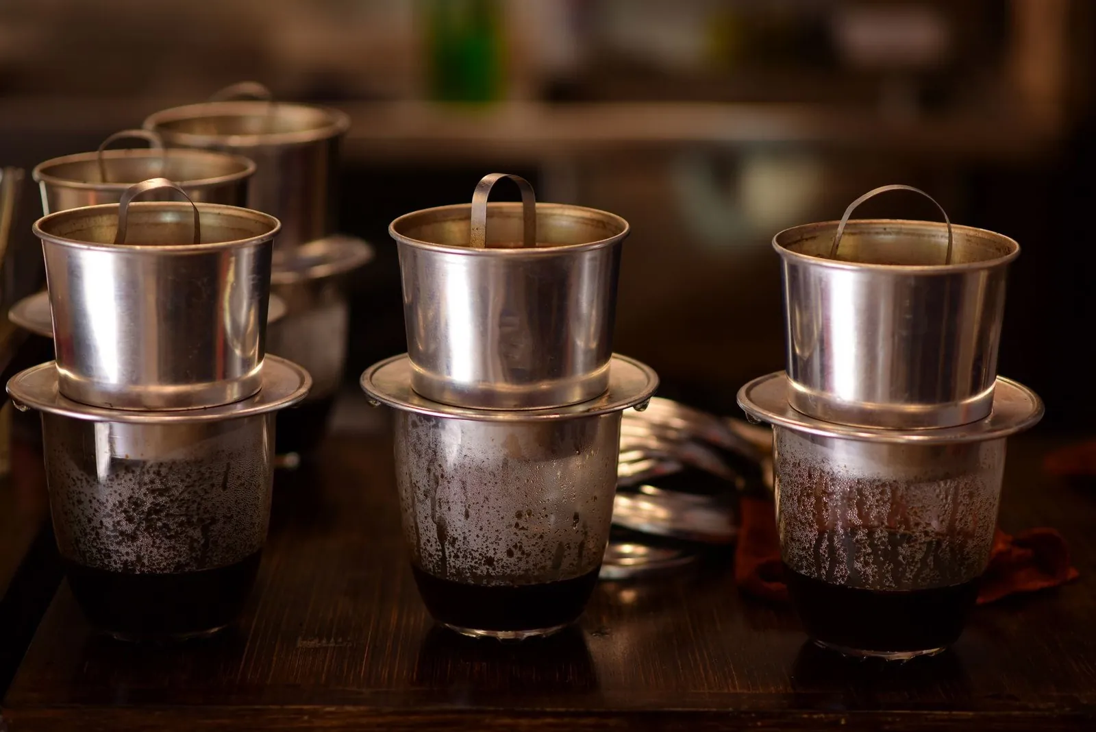 Several metal phin filters actively dripping coffee into glasses on a wooden table